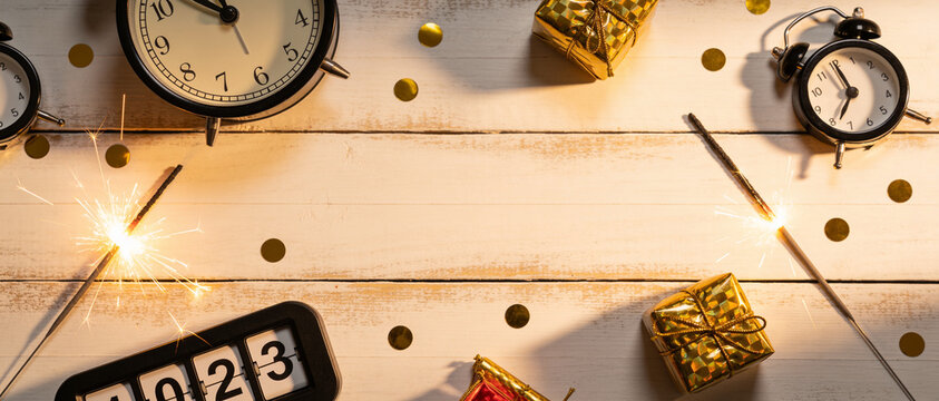 Alarm clocks, gold gift boxes, and a lit sparkler on a wooden surface