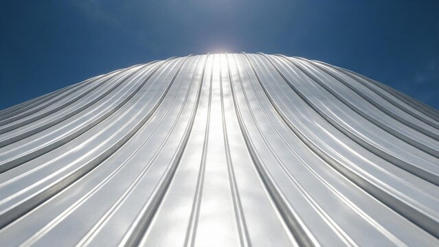 Curved standing seam metal roof with shiny silver aluminium panels rising toward a deep blue sky and sun, low angle abstract architectural detail - Modern metal facade and roofing technology