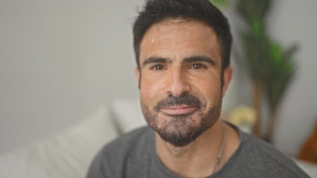 Man smiling with visible teeth and short beard, sitting on bed, wearing gray t shirt, facing camera in studio; contentment ease warmth.