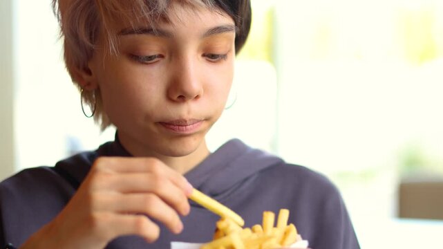 A young girl with colorful hair sits comfortably in a fast-food cafe while eating French fries, presenting a warm, relatable scene of youth, dining, and everyday happiness.