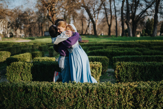 Two women embrace warmly while standing between trimmed boxwood hedges. They hug in a formal garden maze, sharing a joyful outdoor moment.