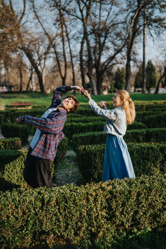 A young couple dance and laugh together while navigating a formal hedge maze in a park. They playfully twirl and enjoy a sunny outdoor moment among trimmed boxwood paths.