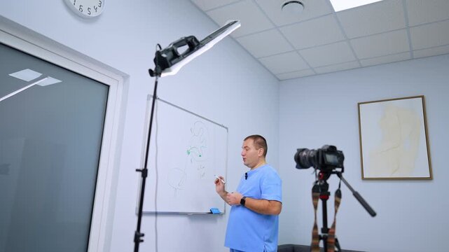 A clinician points toward a whiteboard beside a filming setup. A panel light stands above the camera with a glass door and pale ceiling tiles on the left.