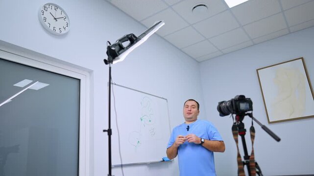 A clinician addresses a camera with his hands raised near his chest. A bright panel lamp stands below a round clock with blue scrubs beside a whiteboard and glass door.