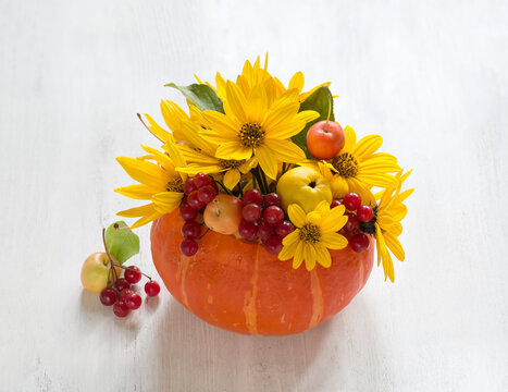 A beautiful autumn arrangement of bright yellow Topinambur flowers,  small Apples, and red Viburnum berries, placed inside an orange pumpkin against a white wooden background.