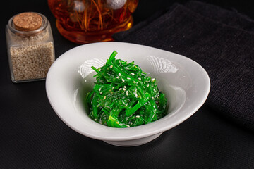 Green chuka seaweed salad with sesame seeds in white ceramic bowl on black table with glass jar and amber dressing bottle background Japanese appetizer © Andrei Starostin