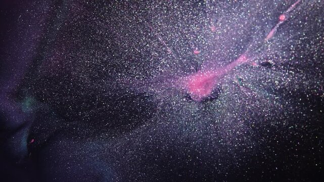 Shimmering glitter pigment bursts through swirling ink, forming a star field style pattern that drifts slowly across frame.