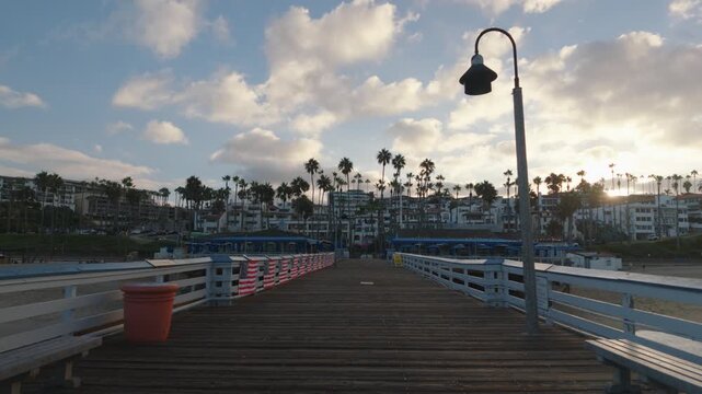 Wide view of the San Clemente Pier with buildings in the background under a partly cloudy sunrise sky and calm waves rolling ashore in Southern California in the soft morning light
