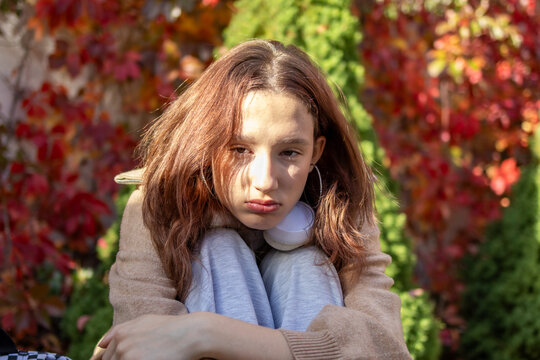 Upset teenage girl with red hair hugs her knees and looks at the camera with a pouting face, headphones around her neck, red ivy behind. Concept of teen burnout, sadness and autumn melancholy.