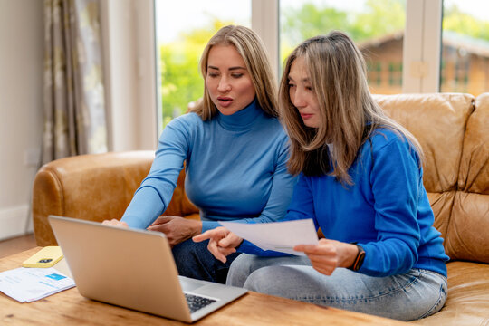 Two women sit on a sofa together while using a laptop and reviewing papers during daytime