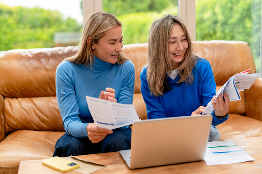 Two women working together at home with a laptop and paperwork on a sofa