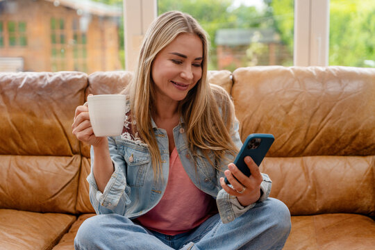Woman sitting on a couch holding a cup while looking at her phone in a living room