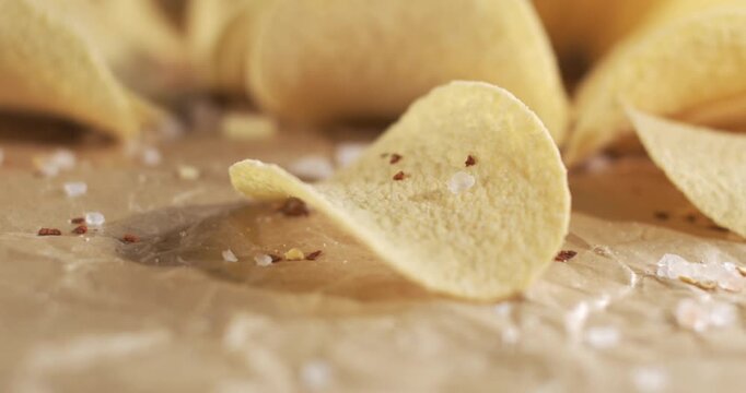 Golden potato chips sit scattered across textured surface, presented for review. Curved crisp stands upright among salted pieces, highlighting their seasoning, color, and appealing texture.