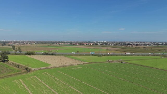 Aerial View of Farmland and Highway: An expansive view of green agricultural fields stretches towards a distant town, bisected by a highway with a line of trucks traveling along it.