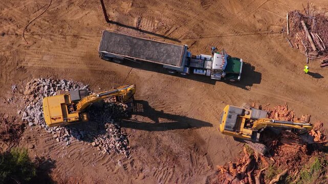 Two yellow excavators work a cleared site: one digs into a gray rock pile, other sits in tree stumps. A dump truck with a full trailer stands on tire tracks as a worker in hi vis walks by in low sun.