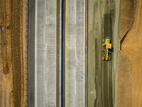 Aerial view of a yellow motor grader working on a new highway construction site with concrete lanes and surrounding earthworks, Salatiga, Central Java, Indonesia.
