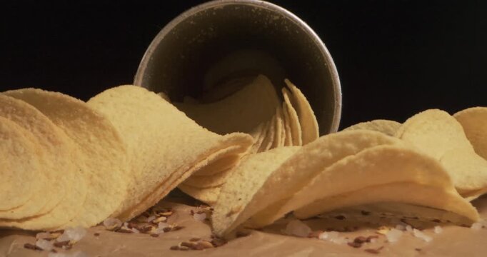 Golden potato chips lie beside open chip box in close view. Crisp snack and loose seasoning remain still against dark background, offering simple appetizing display for viewers to examine.