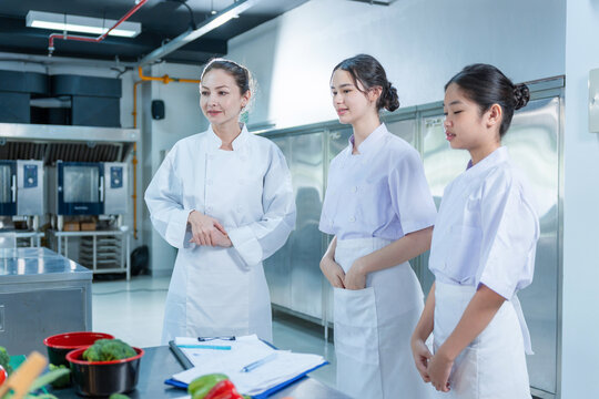 Professional chef giving a lecture to young female students in culinary school training class, Head chef mentor standing with student chefs during training in modern restaurant kitchen