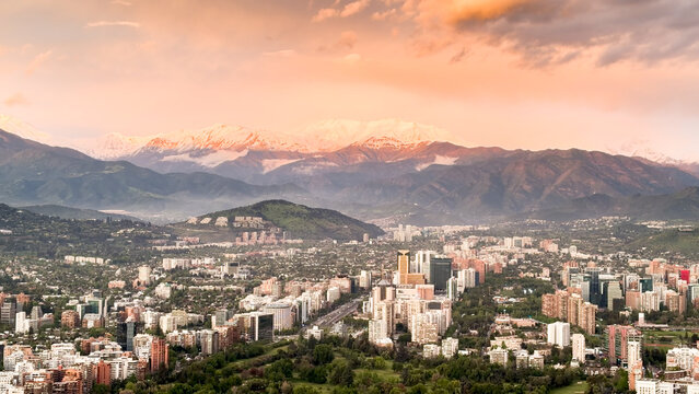 Aerial view of the Santiago skyline with modern skyscrapers, green parks, and snow-capped Andes mountains under a warm sunset sky in Santiago, Santiago Metropolitan, Chile.