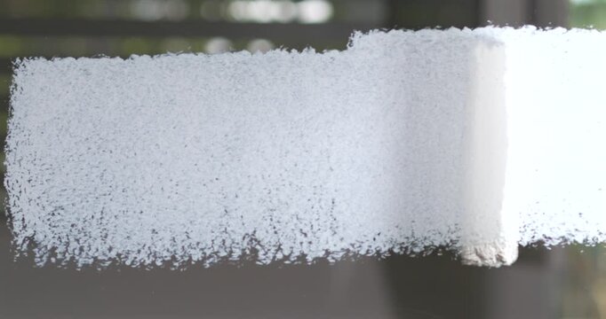 Hand guides brush across glass, spreading thick white coating over surface. Careful painting motion leaves textured, opaque coverage in workshop with dark slatted windows, covering pane with white