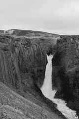 A monochrome view shows Litlanesfoss plunging between basalt columns into a rugged gorge in Fljotsdalur, East Iceland. Mist and foaming rapids define this summer travel landscape. © Artem
