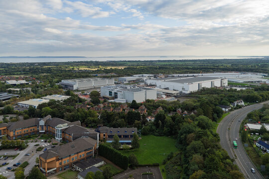 Aerial view of a large data center complex surrounded by lush greenery, office buildings, and a road with a distant sea view in the United Kingdom.