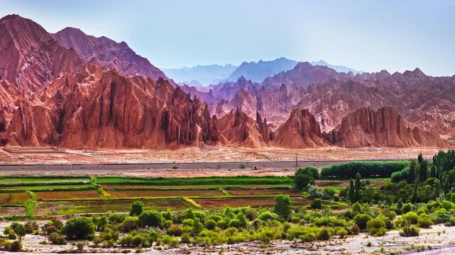 Aerial panorama of a green farmland oasis surrounded by dramatic red sandstone mountain canyons in the Red Mountain Stone Forest, Kuqa, Xinjiang, China