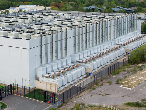 Aerial view of a large data center facility with rows of cooling units and ventilation systems on the roof, surrounded by a security fence and paved grounds in the United Kingdom.