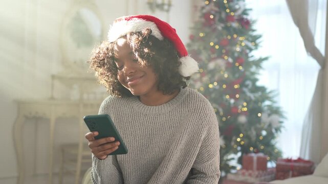 African American woman holding smartphone scrolling typing shopping online buying Christmas gifts. Girl buying on Internet chatting in social network enjoying Christmas winter holidays at home