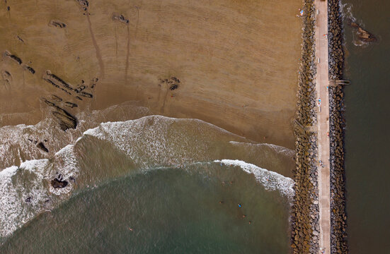 Abstract aerial top-down texture of sandy beach, sea waves, and stone breakwater jetty in Cand&aacute;s, Asturias, Spain.