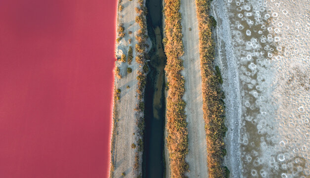 Aerial view of a pink salt evaporation pond separated from a dark water channel and dry mineral-covered ground by parallel embankments in Aigues-Mortes, Occitanie, France.