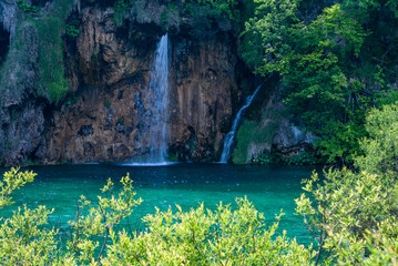 Crystal-clear turquoise water flows gently in front of rocky cliff, where graceful waterfall cascades down, surrounded by lush green vegetation and trees. Plitvice Lakes is National Park in Croatia. © MarinoDenisenko