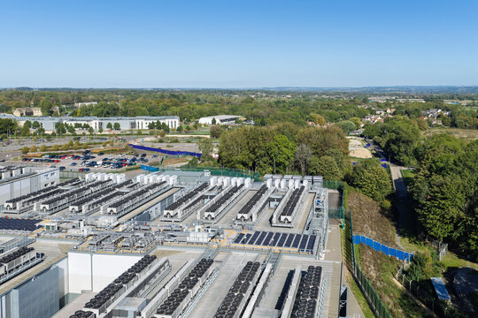 Aerial view of a large data center facility featuring rows of cooling units on the roof, surrounded by lush green trees and a parking area in the United Kingdom.