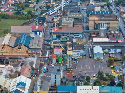Aerial view of an industrial factory complex featuring production buildings, a water tower, railway tracks with freight cars, and smoking chimneys in Thann, Grand Est, France.