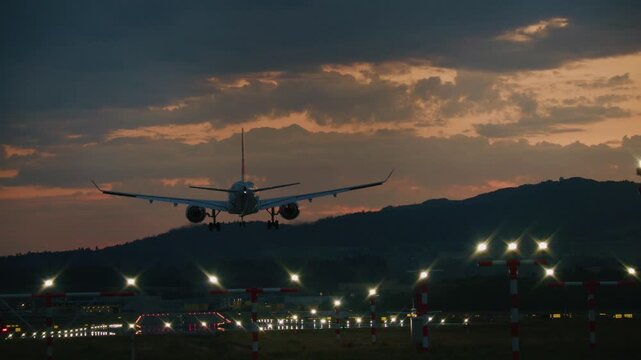 Airport runway and apron illuminated by taxiway light rows with distant aircraft silhouettes under dramatic sunset cloud sky. Airplane lands on runway. Jet airliner landing on runway