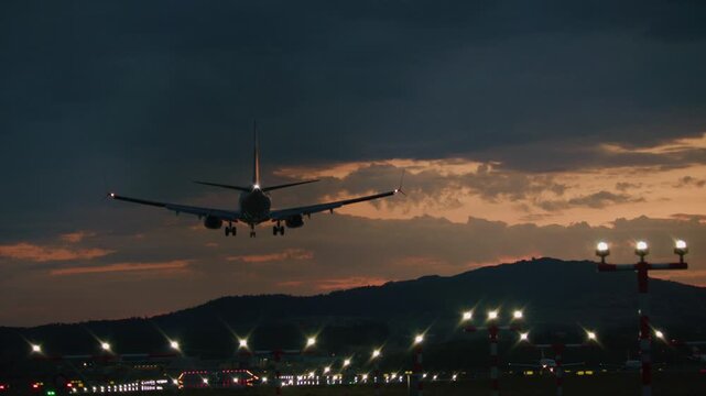 Dusk airport runway and apron illuminated by taxiway light rows with distant aircraft silhouettes under dramatic sunset cloud sky
