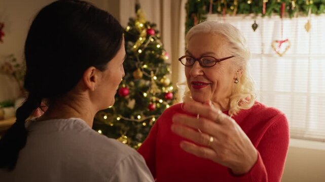 Caucasian senior woman talking and embracing daughter near Christmas tree during holiday visit indoors. Useful for eldercare services, family connection, and seasonal marketing