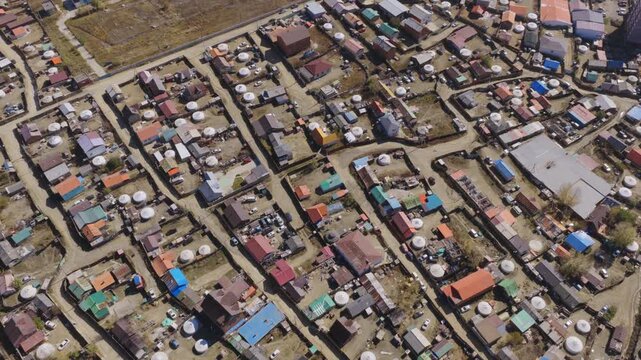 Top-down drone shot of a Ulaanbaatar ger district, showing white felt gers and small houses with colorful roofs inside fenced yards, separated by dusty unpaved streets.