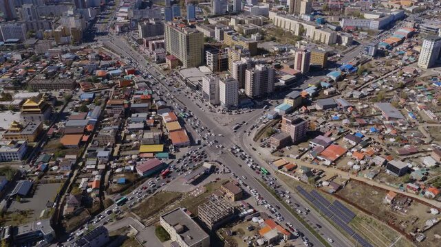 Aerial shot of a busy Ulaanbaatar intersection packed with traffic, with golden temple roofs on the left, new high-rises behind, and solar panels and gers on the right.