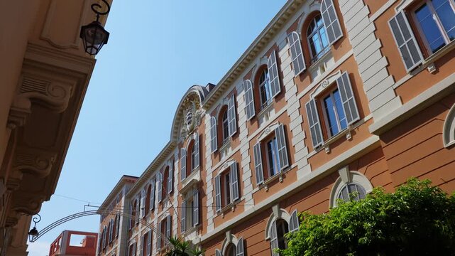 Upward view along an orange historic building in Monaco-Ville with grey louvered shutters, white stone trim and an iron lantern bracket against a blue summer sky. Low angle shot