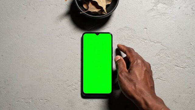 Hand Grips and Swipes Green Screen Smartphone on Textured Gray Surface with Dried Leaves in Black Bowl