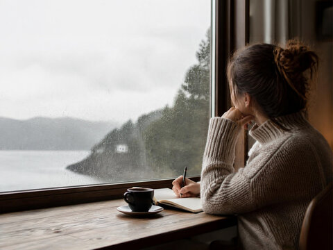Young person journaling by a rainy window, mental wellbeing concept