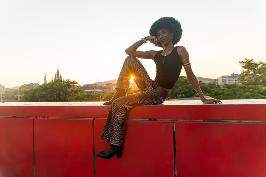 Stylish queer man sits on a red wall outdoors in Barcelona at sunset