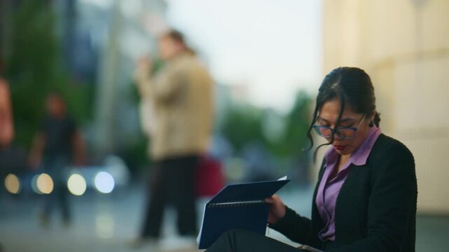 An Asian businesswoman in glasses reviews documents while sitting on a city street. She takes a moment to pause and reflect amidst the urban bustle.