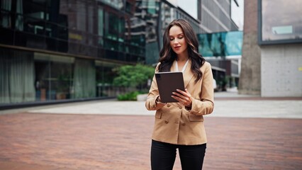 A focused businesswoman stands in a paved urban plaza while reviewing information on her handheld device near contemporary glass architecture © VAKSMANV