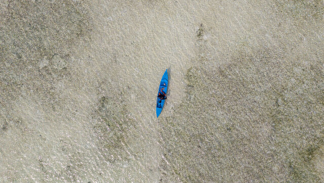 Aerial view of a person paddling a blue kayak through the shallow, crystal-clear turquoise waters of a coral reef in Wakatobi, Southeast Sulawesi, Indonesia.