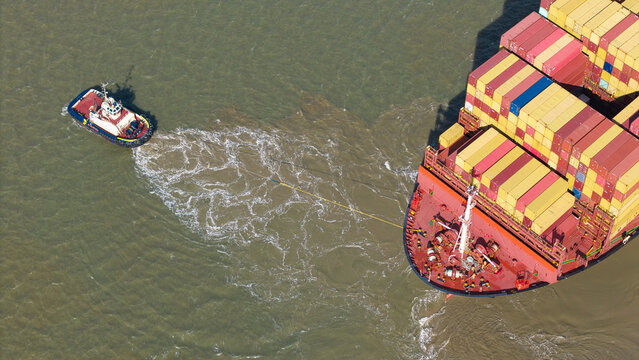 Aerial view of a tugboat maneuvering a large container ship loaded with colorful shipping containers in the Port of Felixstowe, Felixstowe, England, United Kingdom.
