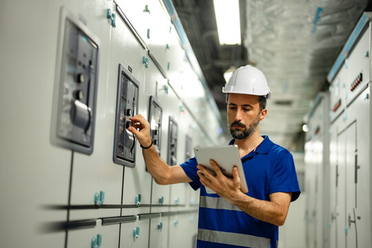 Male technician adjusts electrical switchgear controls while checking digital tablet in hospital control room. Equipment inspection, power system maintenance, technical monitoring, facility safety.