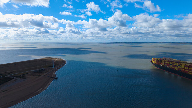Aerial view of a container ship navigating near the Port of Felixstowe with a coastal spit and lighthouse visible under a blue sky Felixstowe, England, United Kingdom.