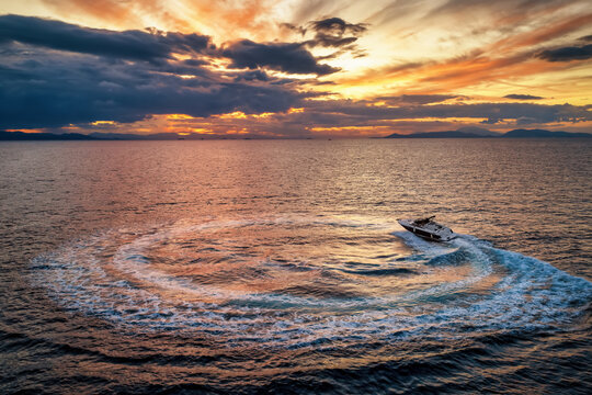 Aerial view of a motorboat forming a circle of waves and bubbles over the ocean during a dramatic sunset with golden light